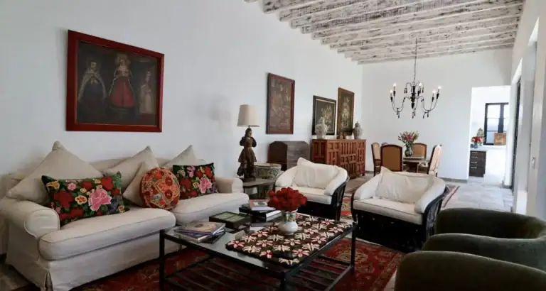 Bright living room with white sofa and floral cushions, a glass coffee table on a red patterned rug, and a chandelier visible in the dining area, San Miguel de Allende