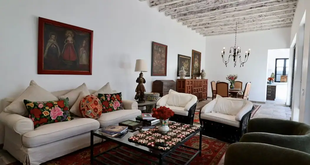Bright living room with white sofa and floral cushions, a glass coffee table on a red patterned rug, and a chandelier visible in the dining area, San Miguel de Allende