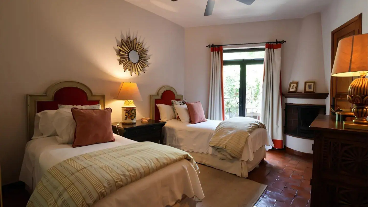 Twin-bed hotel room with a sunburst mirror, two lamps on a dark wood nightstand, and white bedding with pink pillows, San Miguel de Allende