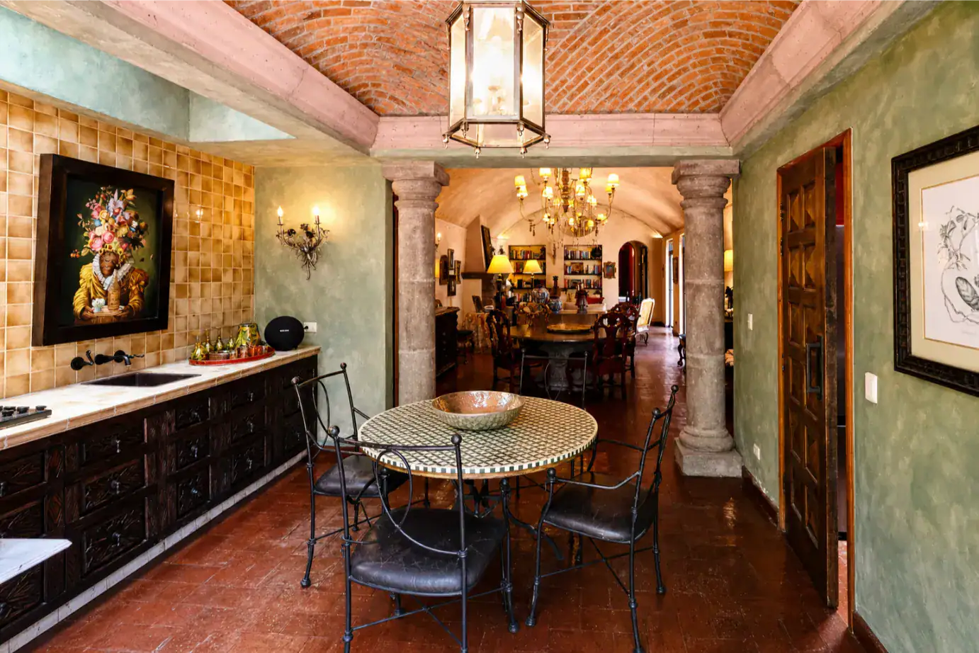 Round mosaic dining table with black metal chairs in a rustic, columned room; brick vaulted ceiling and chandelier create an elegant, open-kitchen feel, San Miguel de Allende