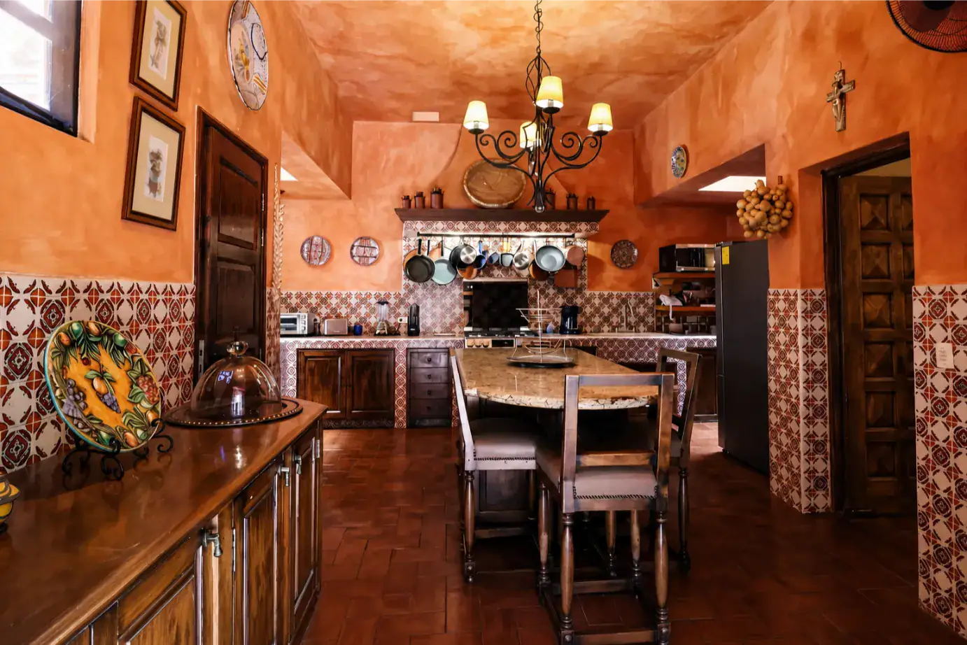 Rustic Mediterranean-style kitchen with orange plaster walls, patterned tile wainscoting, a marble-topped central island, and wooden chairs around it under a wrought-iron chandelier, San Miguel de Allende