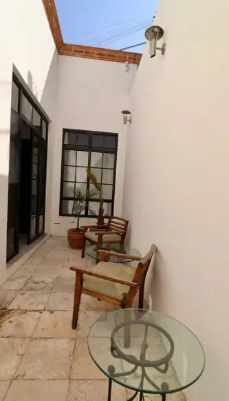 Narrow white-walled courtyard with wooden chairs around a glass circular table, potted plants by a large window, blue sky above, San Miguel de Allende