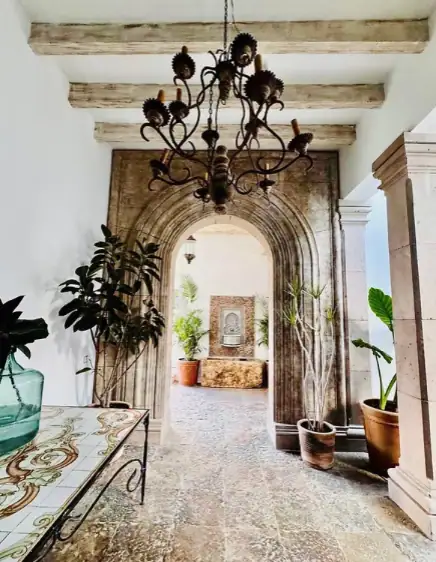 Stone arched doorway with an ornate chandelier overhead; potted plants line a tiled hallway leading to a sunlit room, San Miguel de Allende