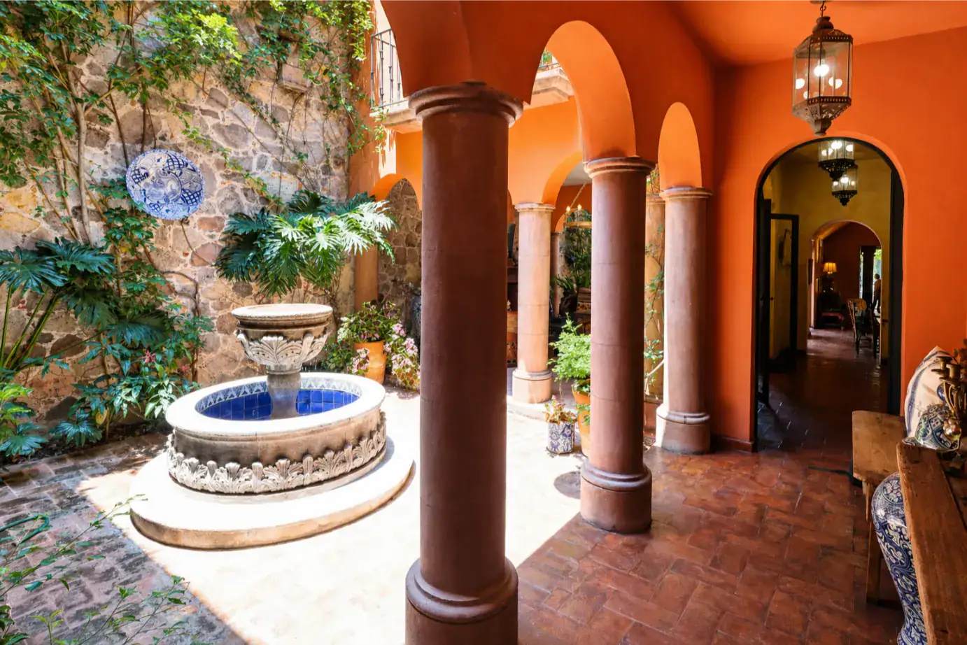 Sunlit orange-walled courtyard with rounded arches, brown columns, a stone fountain, and lush potted plants, San Miguel de Allende