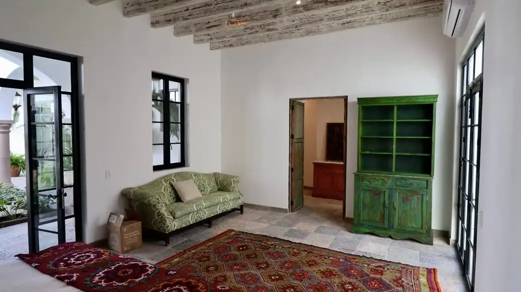 Bright living room with white walls, exposed wooden beams, a green vintage cabinet, a green patterned sofa, and a large red oriental rug; doorway to another room visible, San Miguel de Allende