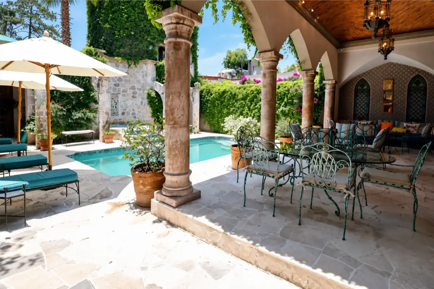 Outdoor poolside patio with stone columns, arches, and wrought-iron seating under umbrellas on a sunny day, San Miguel de Allende