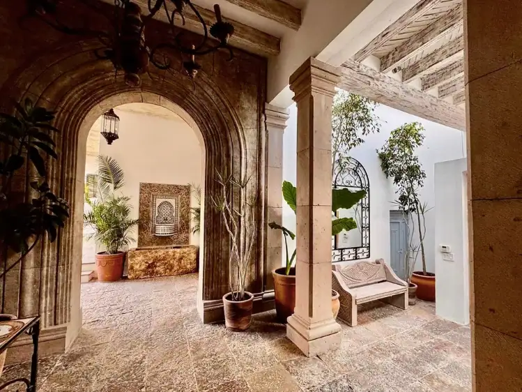 Stone archway leads into a sunlit courtyard with potted plants, a bench, and a wrought iron trellis, San Miguel de Allende