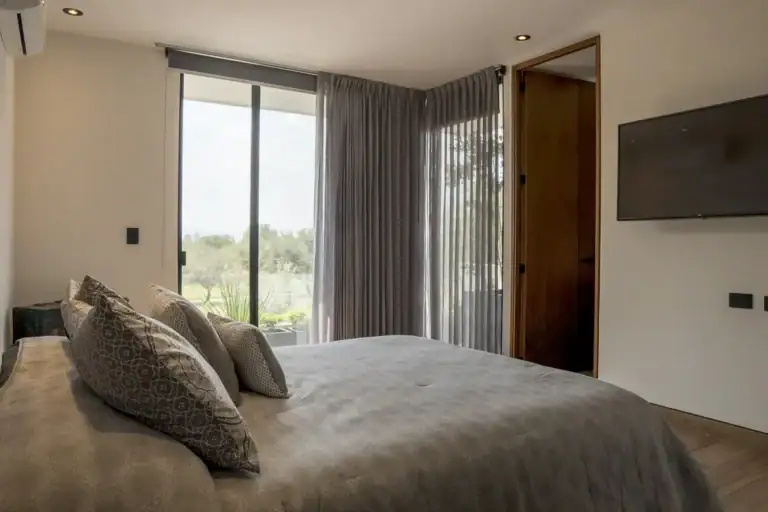 Bright House, bedroom with a gray bedspread, a wall-mounted television and a corner window with sheer curtains, San Miguel de Allende