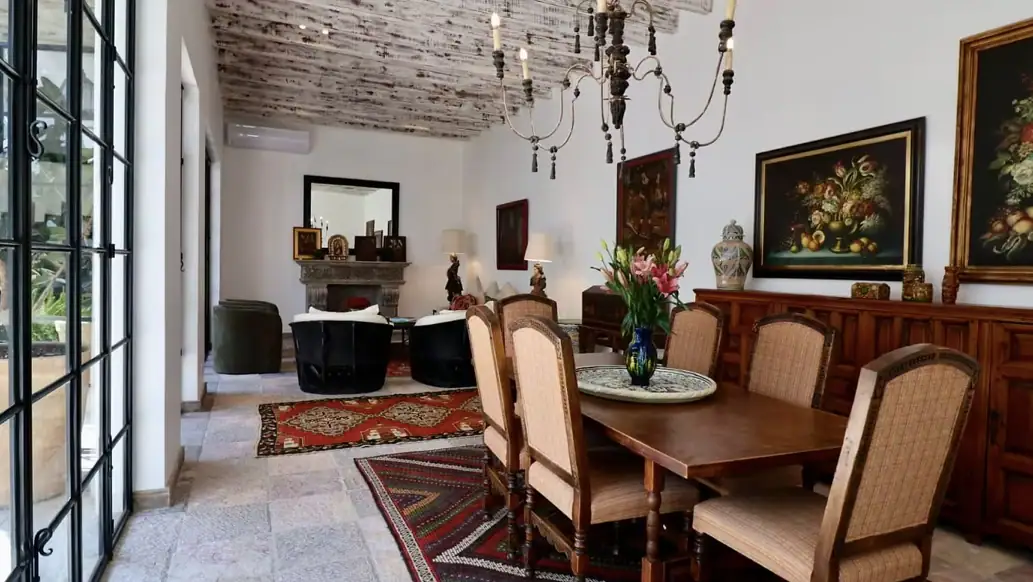 Formal dining room with a dark wood table, beige upholstered chairs, and a chandelier; patterned rugs and a sideboard with paintings along the wall, San Miguel de Allende