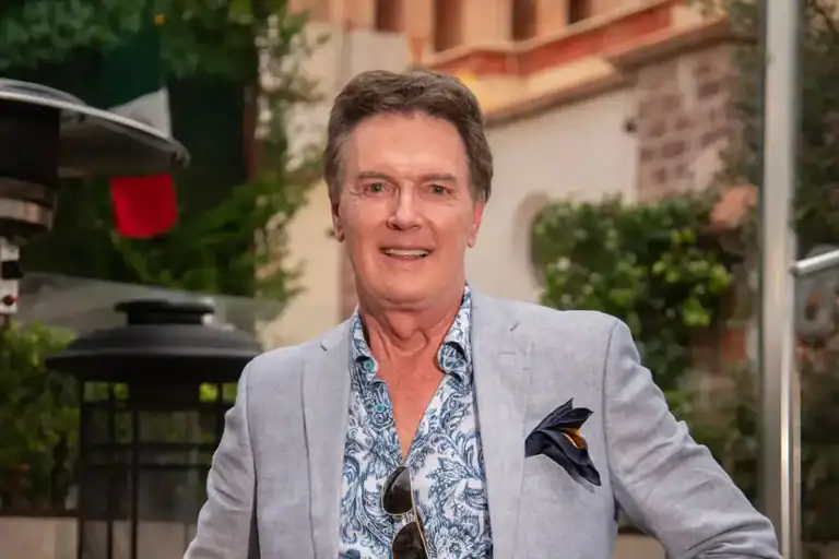 Greg Gunter in a light blue blazer and blue floral shirt standing on a terrace with a stone church tower behind him