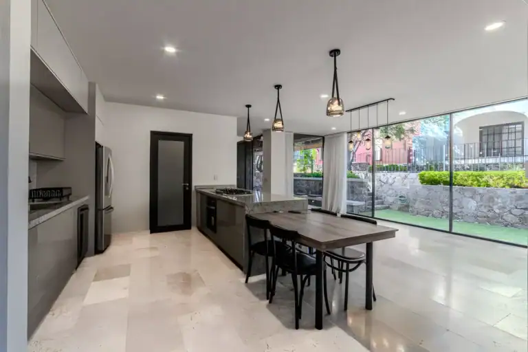Casa Albero, kitchen with a long island with a gas cooktop, a wood dining table, black chairs and a glass wall to the lawn, San Miguel de Allende