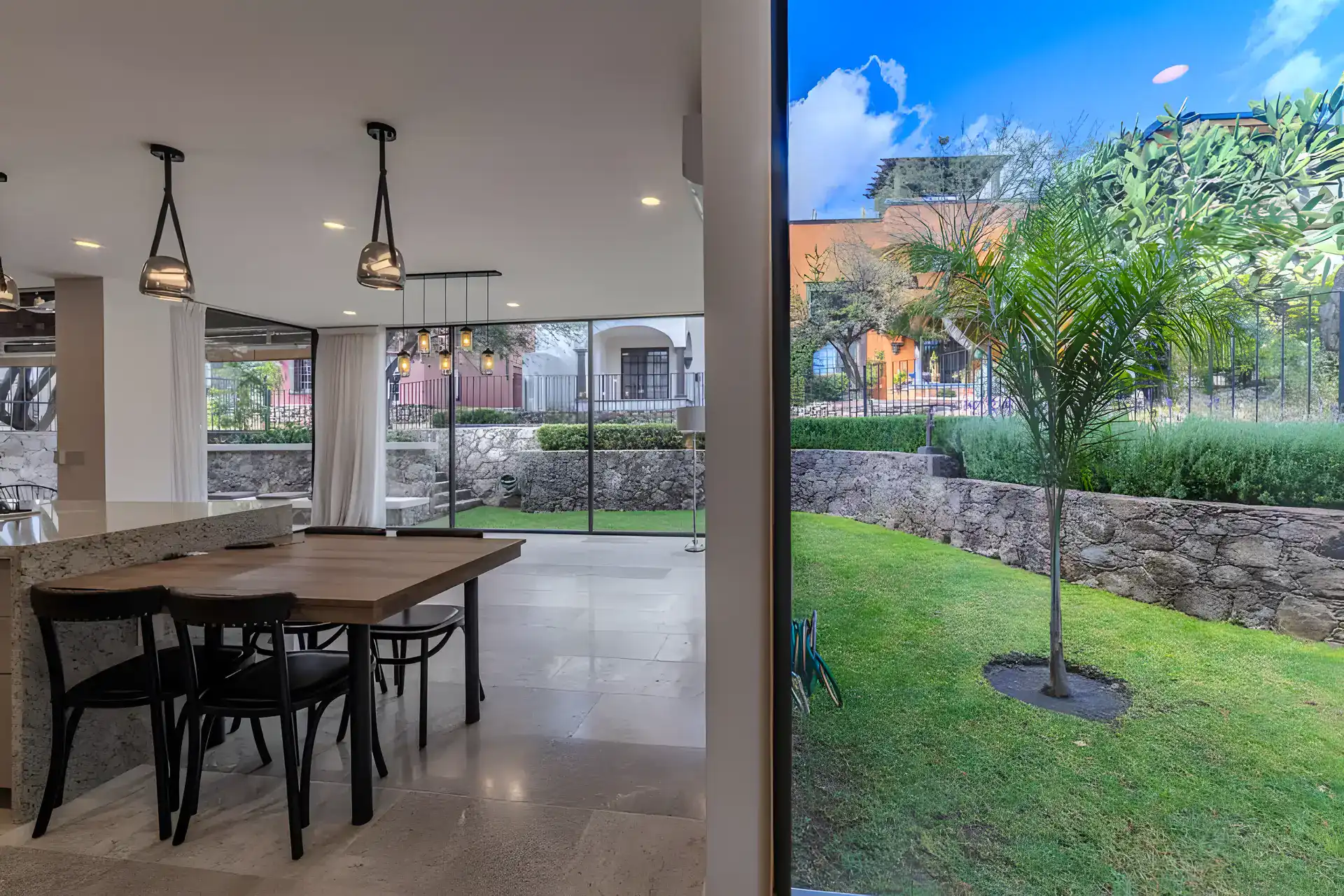 Casa Albero, dining table and glass wall opening to a garden lawn with a young tree and stone retaining wall, San Miguel de Allende