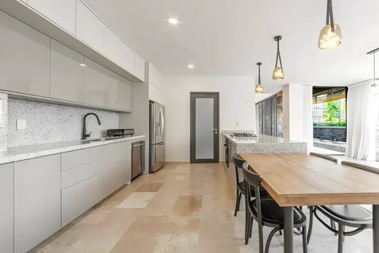 Casa Albero, kitchen with white handleless cabinets, a speckled stone backsplash and a stainless refrigerator beside the dining table, San Miguel de Allende