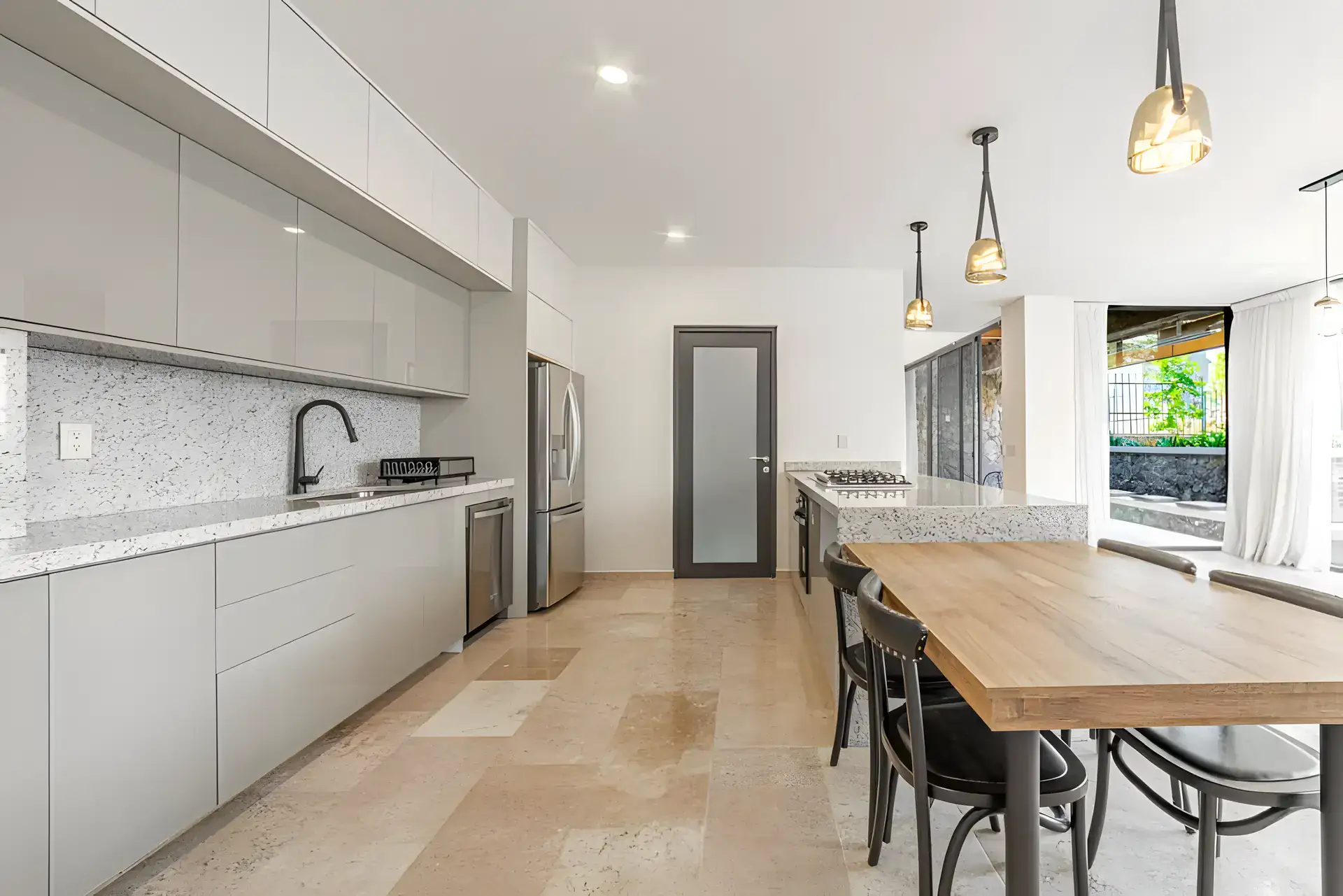 Casa Albero, kitchen with white handleless cabinets, a speckled stone backsplash and a stainless refrigerator beside the dining table, San Miguel de Allende
