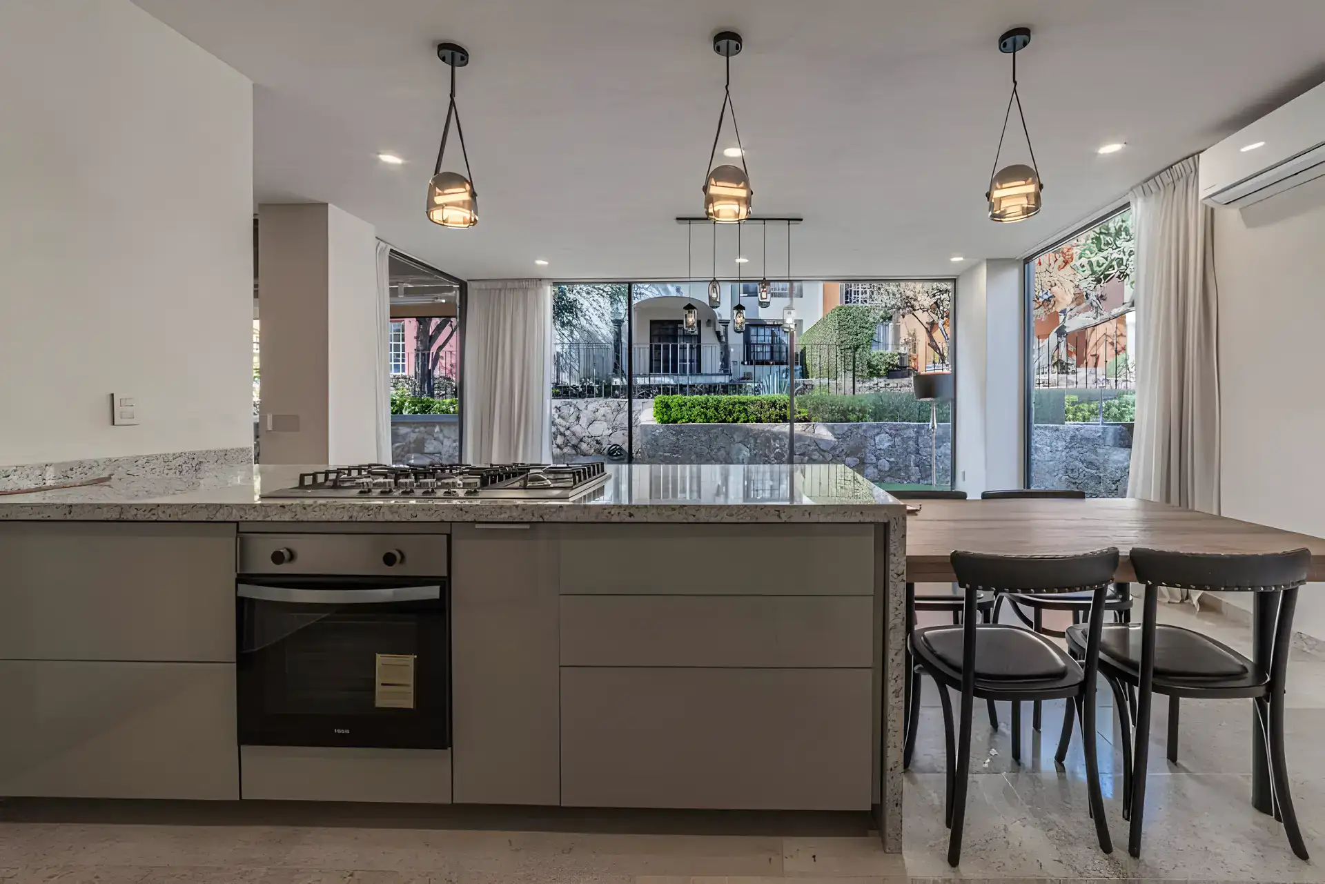 Casa Albero, kitchen island with a built-in oven and cooktop and pendant lights facing a garden wall of glass, San Miguel de Allende