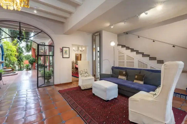 Casa Aldama, living room with a blue sofa, white slipcovered chairs, a red rug and a staircase beyond a steel-framed glass door, San Miguel de Allende