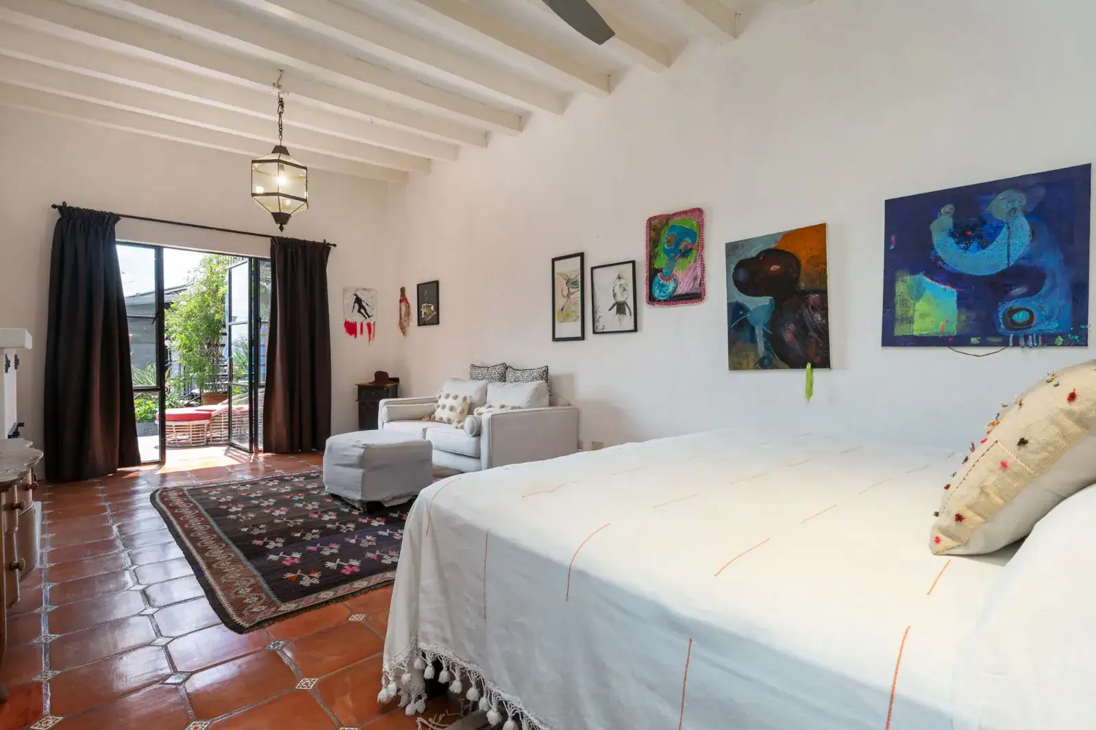 Casa Aldama, bedroom with a white bed, an armchair, colorful paintings and a terrace door, San Miguel de Allende