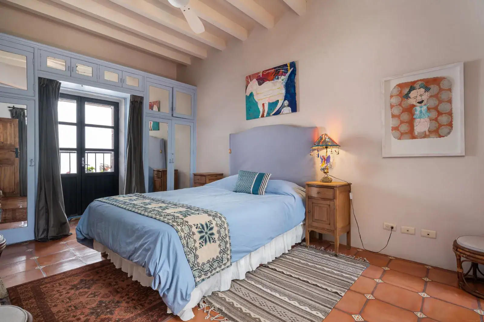 Casa Aldama, bedroom with a blue bedspread, a blue headboard, paintings and a glass-paned door, San Miguel de Allende