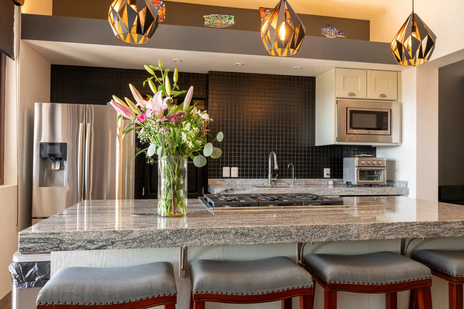 Casa Atardecer, kitchen island with a granite counter, bar stools, a gas cooktop and a vase of pink lilies under geometric pendant lights, San Miguel de Allende