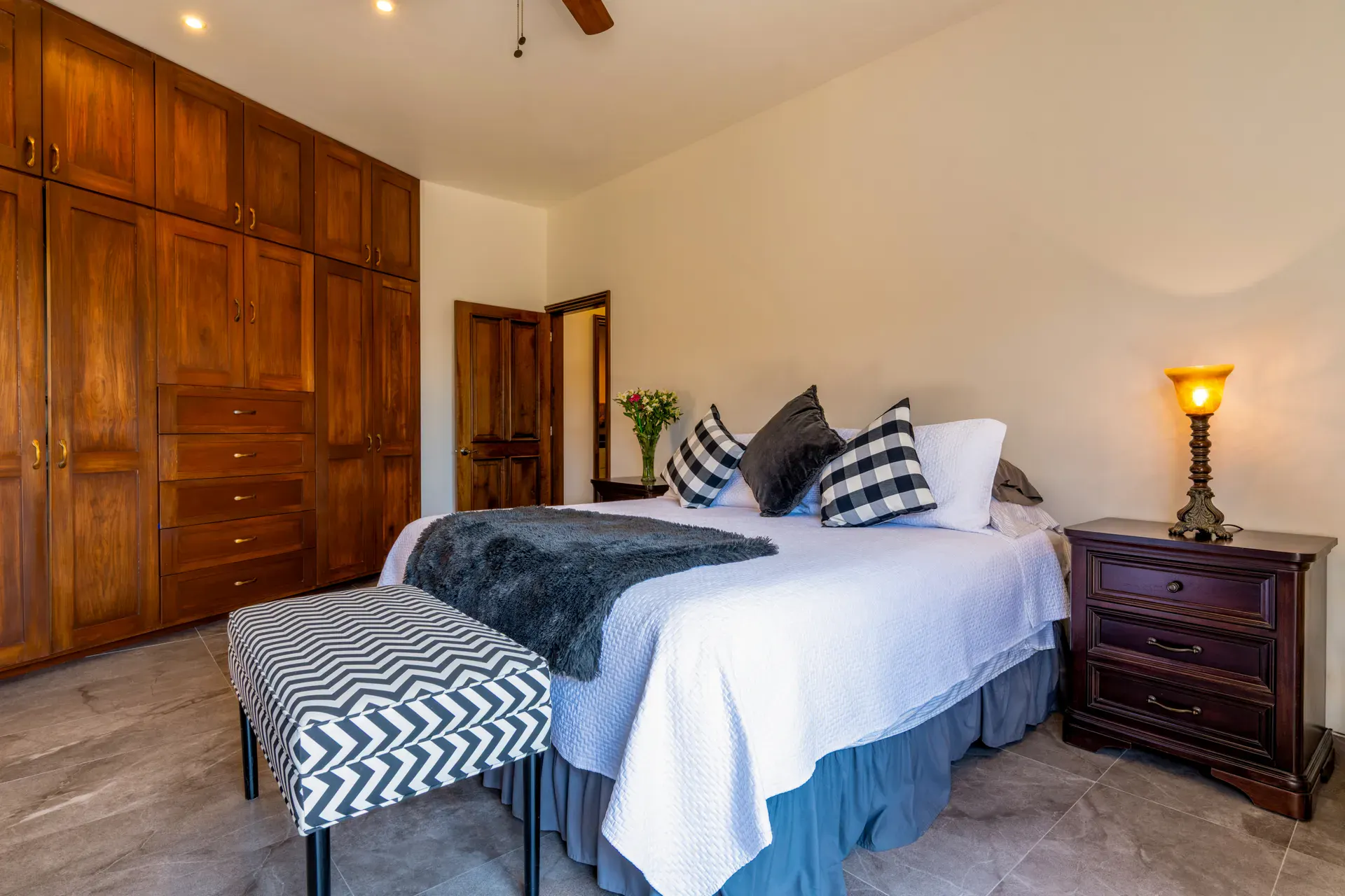 Casa Atardecer, bedroom with a white bed, a plaid bench, a wall of wood wardrobes and a lamp on a nightstand, San Miguel de Allende