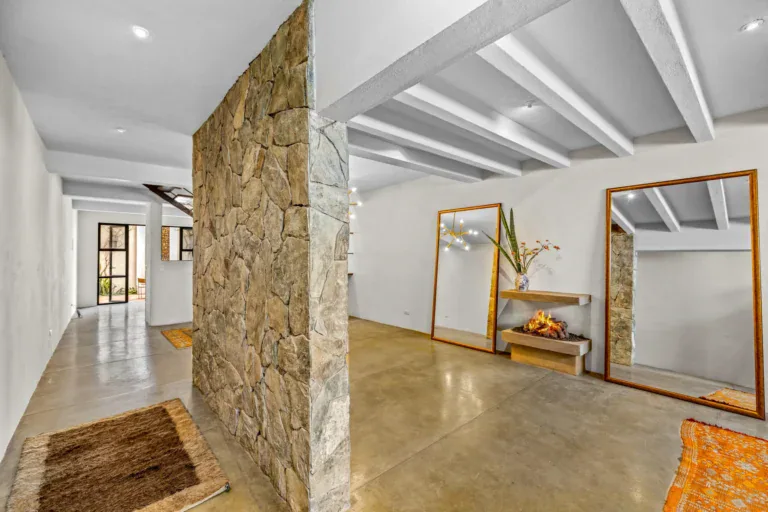 Casa Beatrice, entry hall with a rough stone wall, a long concrete floor and a console with a lit fireplace and tall mirrors, under white beams, San Miguel de Allende