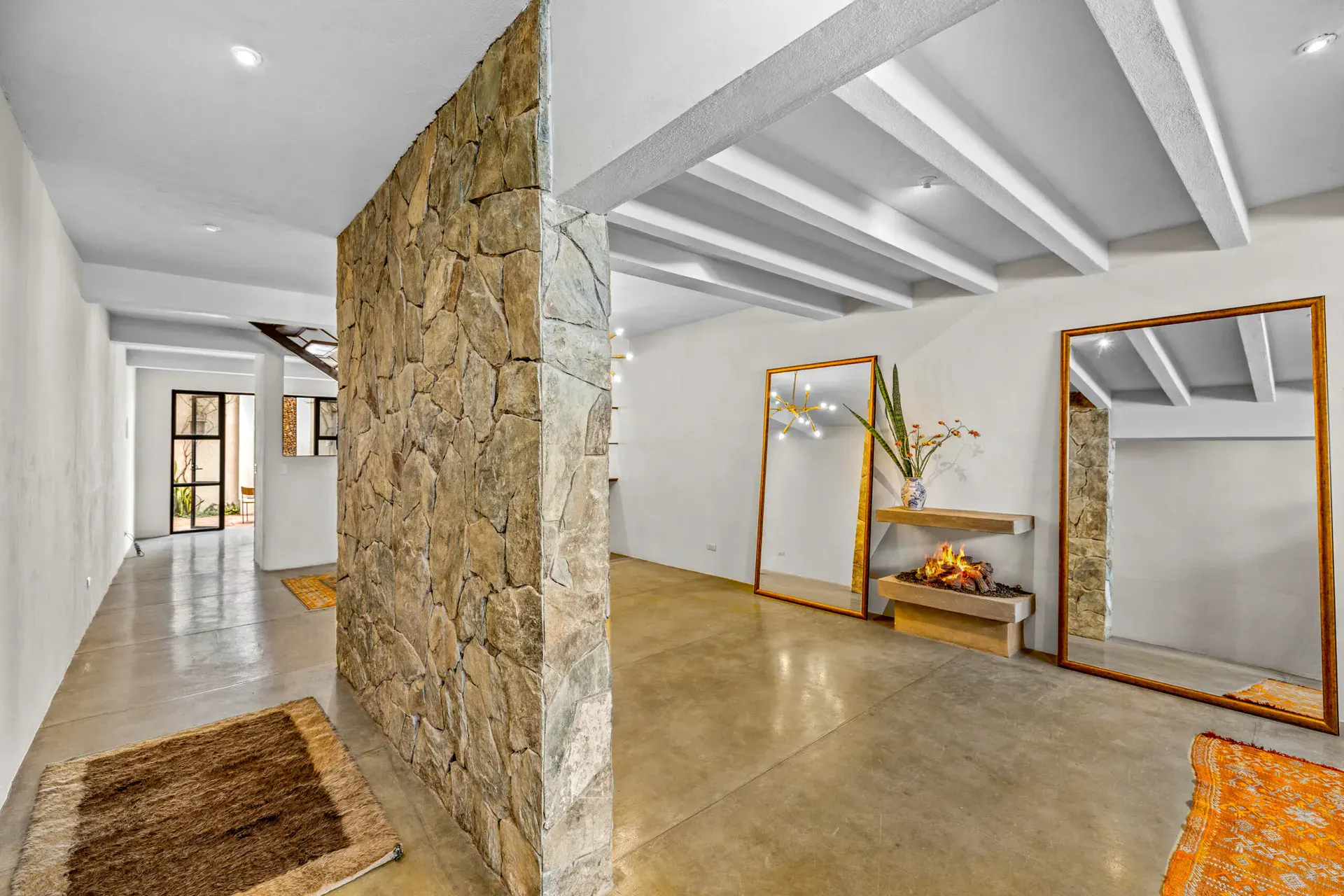 Casa Beatrice, entry hall with a rough stone wall, a long concrete floor and a console with a lit fireplace and tall mirrors, under white beams, San Miguel de Allende