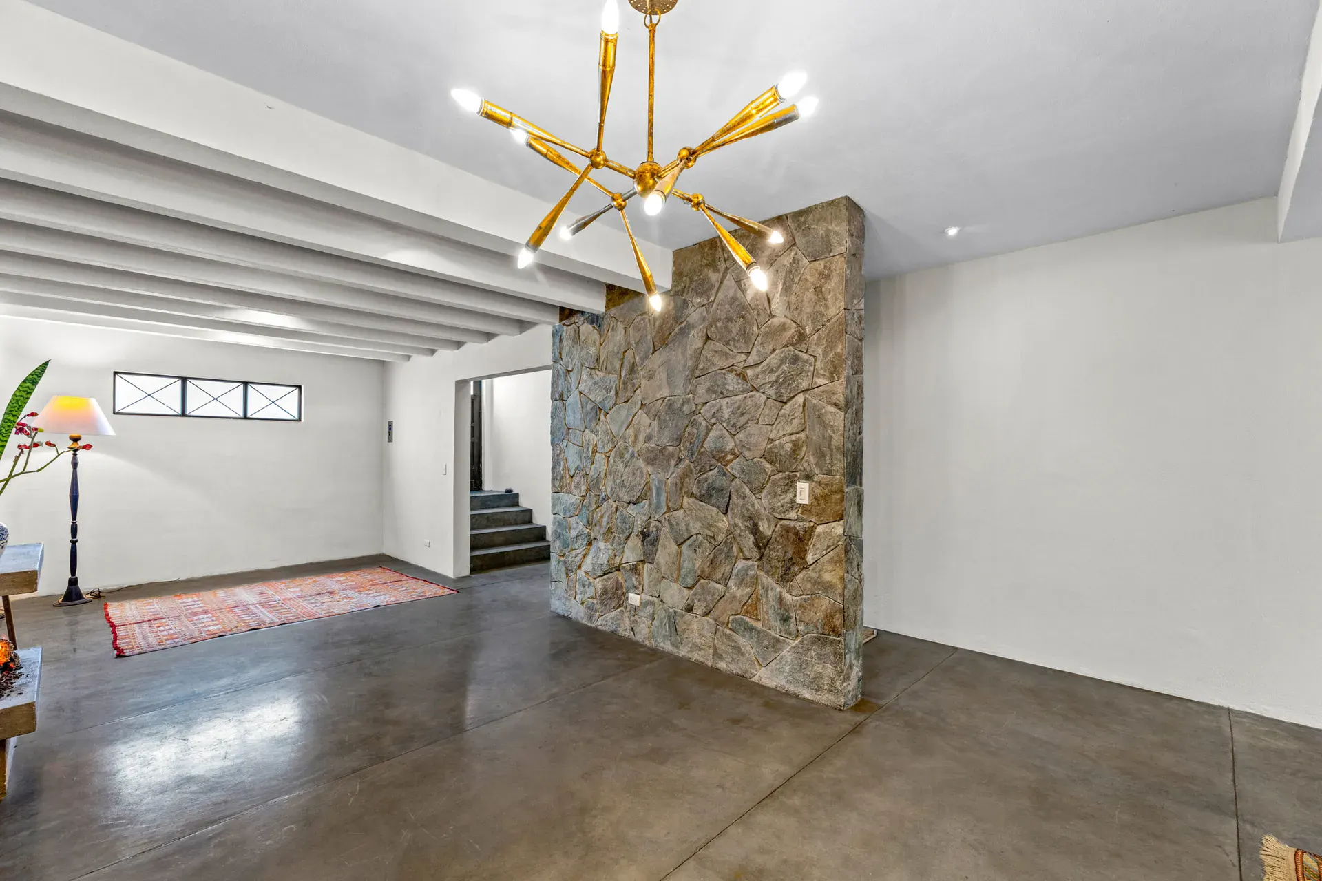 Casa Beatrice, empty living area with a gold sputnik chandelier, a stone feature wall, white beams and a stair, San Miguel de Allende
