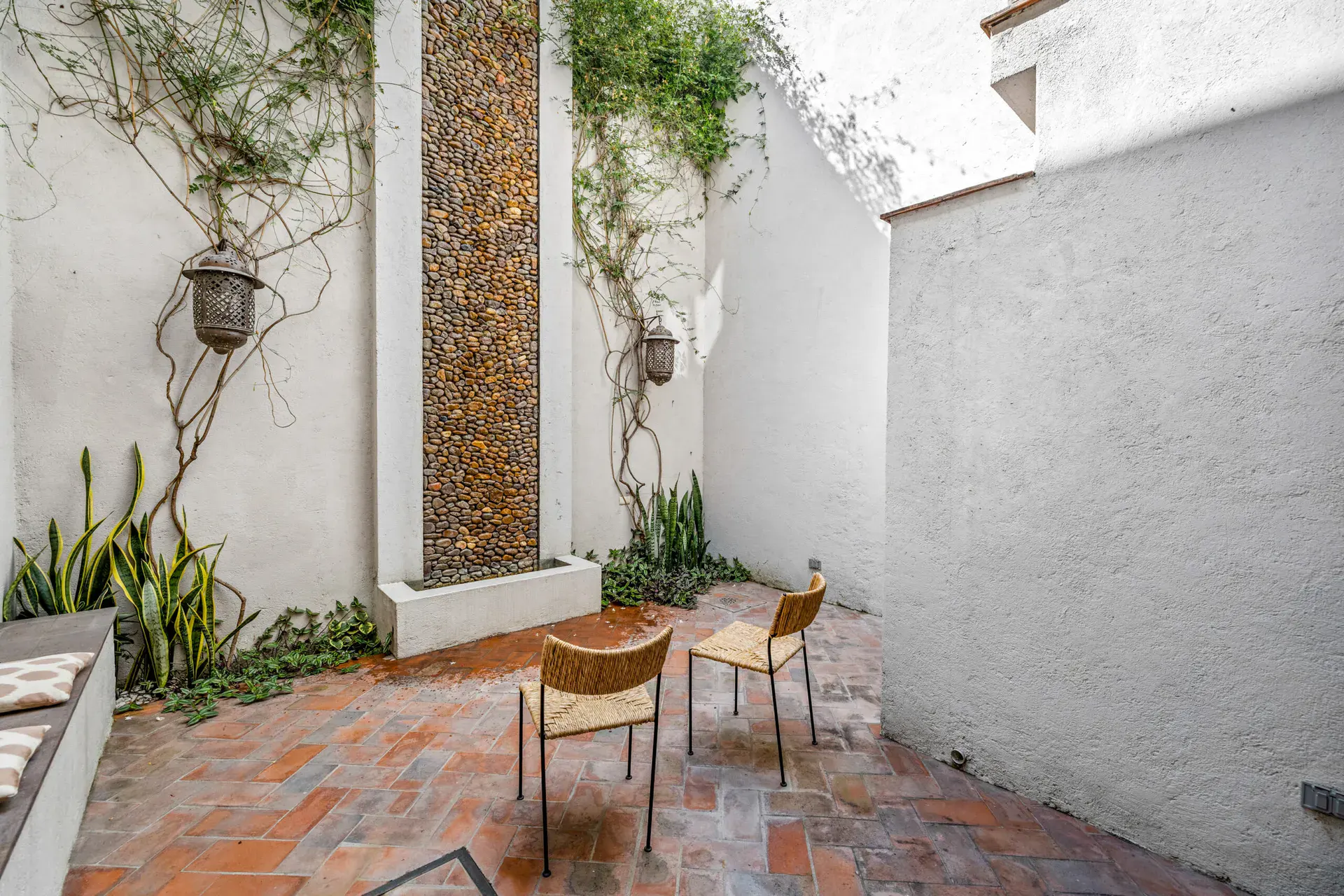 Casa Beatrice, courtyard with a herringbone terracotta floor, a vertical pebble fountain wall, climbing vines and two woven chairs, San Miguel de Allende