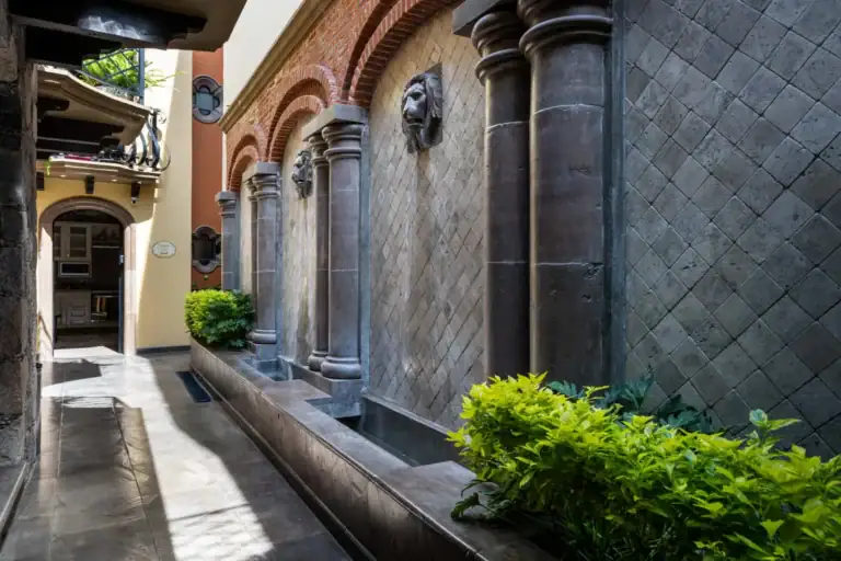 Casa Biblioteca, covered walkway lined with stone columns, a tiled wall fountain and green plants, San Miguel de Allende