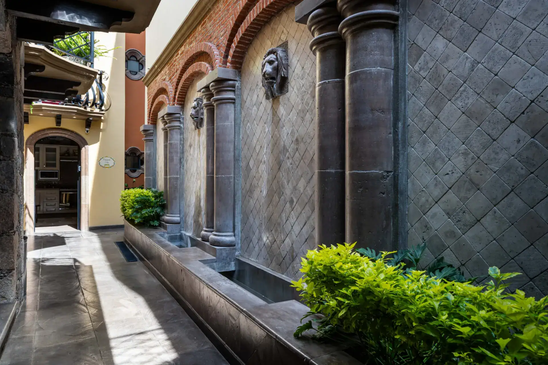 Casa Biblioteca, covered walkway lined with stone columns, a tiled wall fountain and green plants, San Miguel de Allende