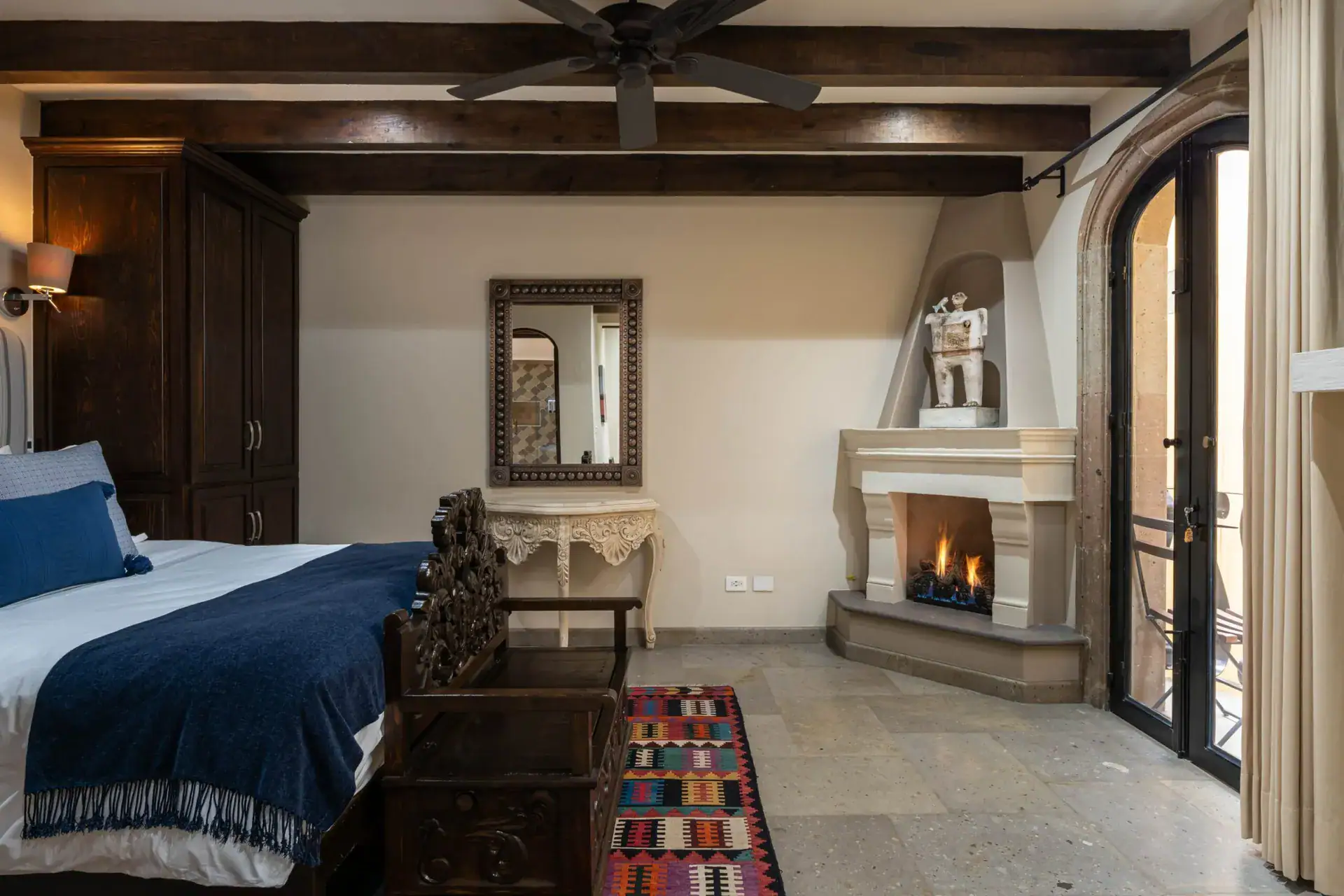 Casa Biblioteca, bedroom with a navy bed, corner fireplace and a patterned rug beside a balcony door, San Miguel de Allende