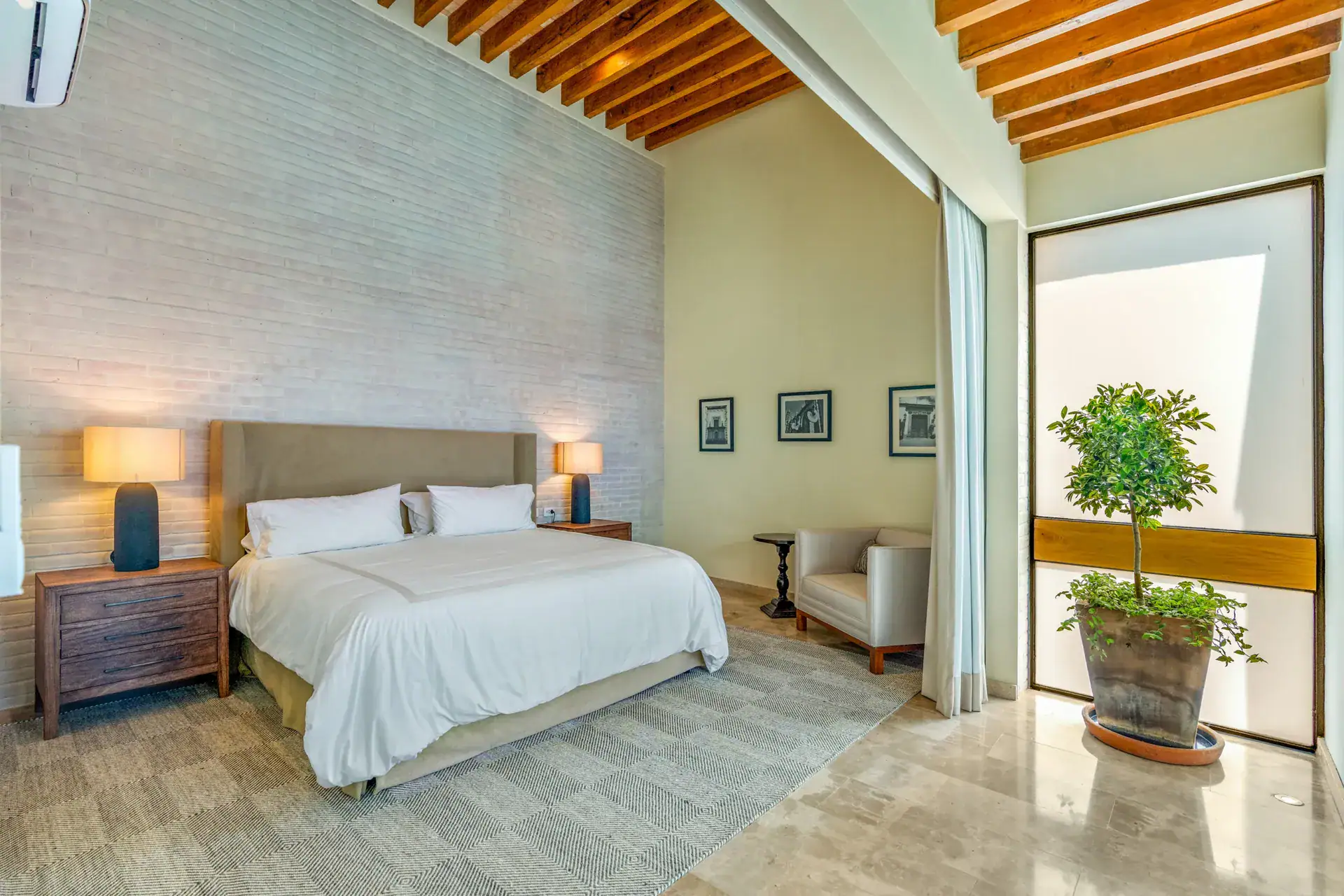 Casa Buenos Tiempos, bedroom with a tan upholstered headboard, a brick feature wall and a potted tree by a glass door, San Miguel de Allende
