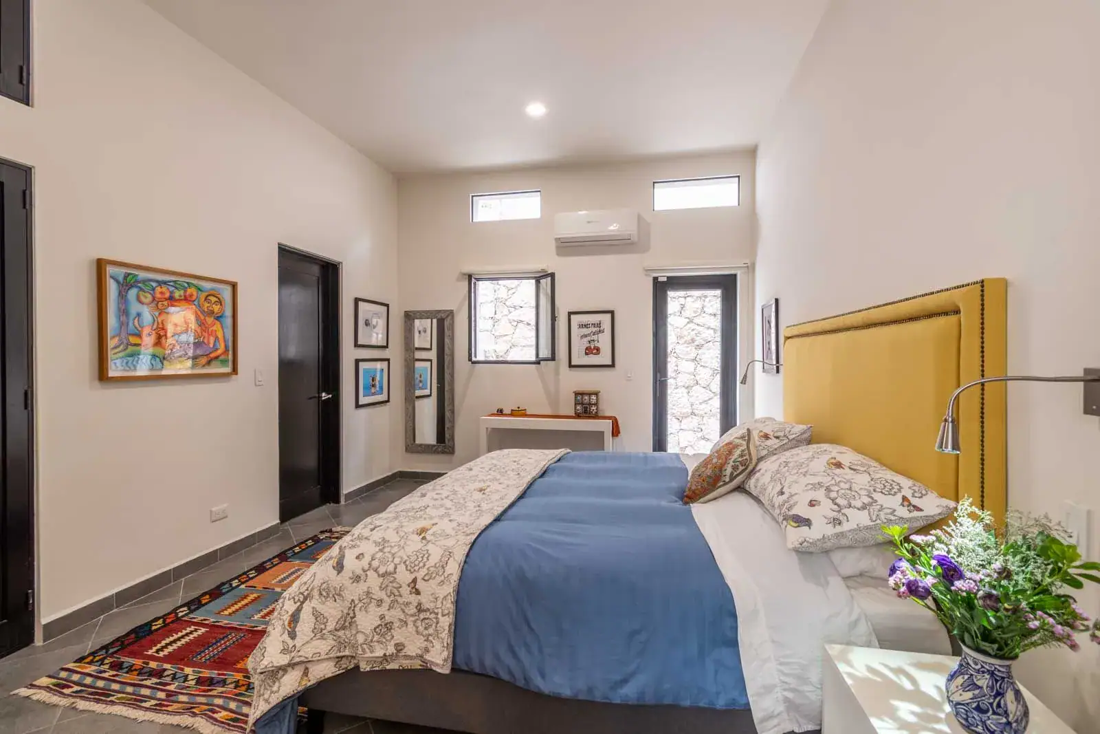 Casa Cielo Abierto, bedroom with a yellow headboard, a blue bedspread, framed art and a patterned rug, San Miguel de Allende