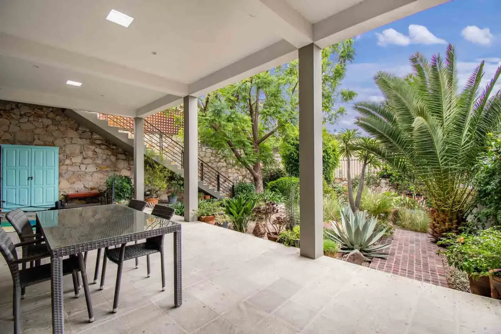 Casa Cielo Abierto, covered patio with a glass table and chairs, a stone retaining wall and a view of palms, San Miguel de Allende