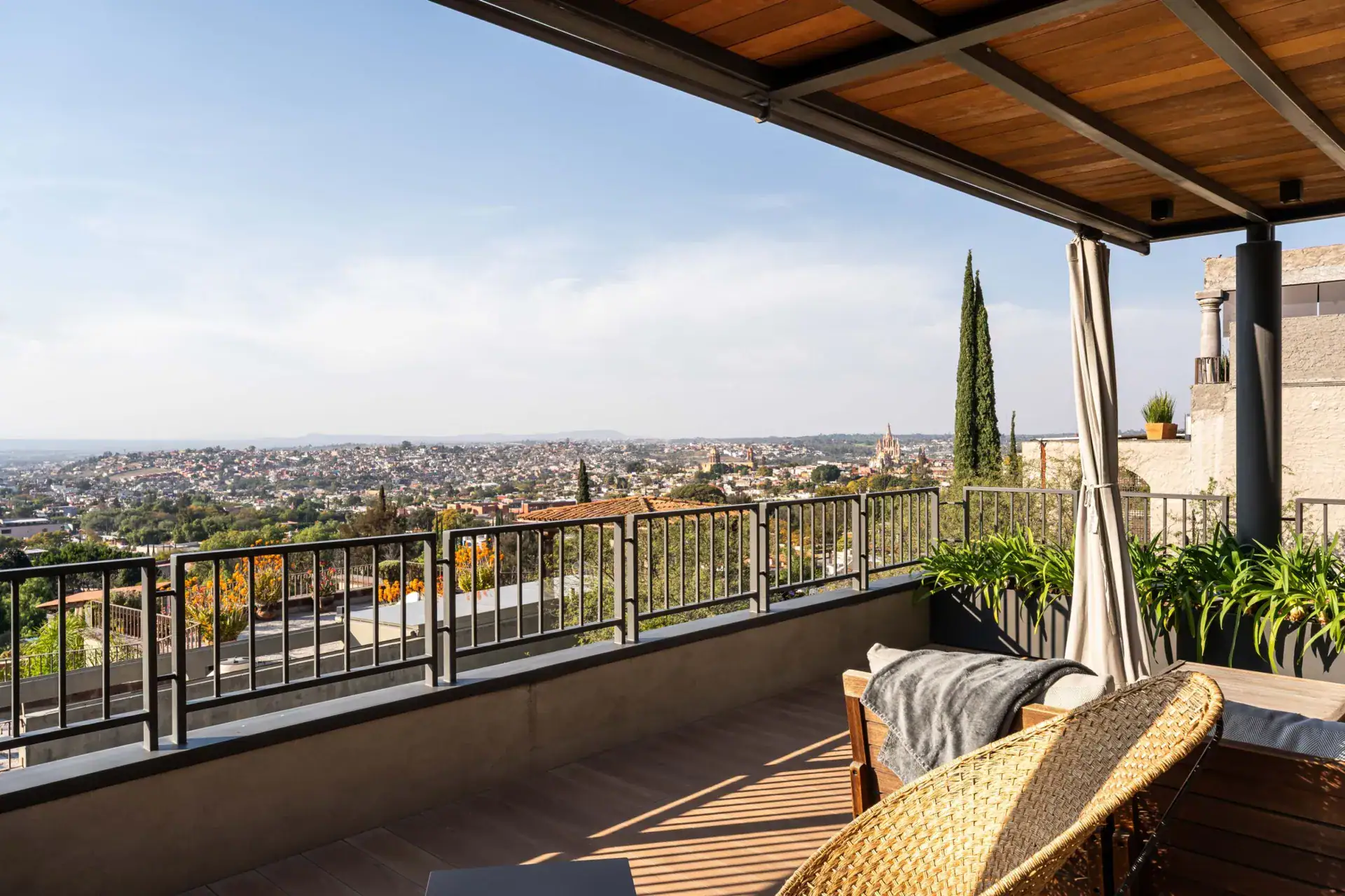 Casa Core, rooftop terrace corner with woven chairs, a sofa and a curtained view to the valley, San Miguel de Allende