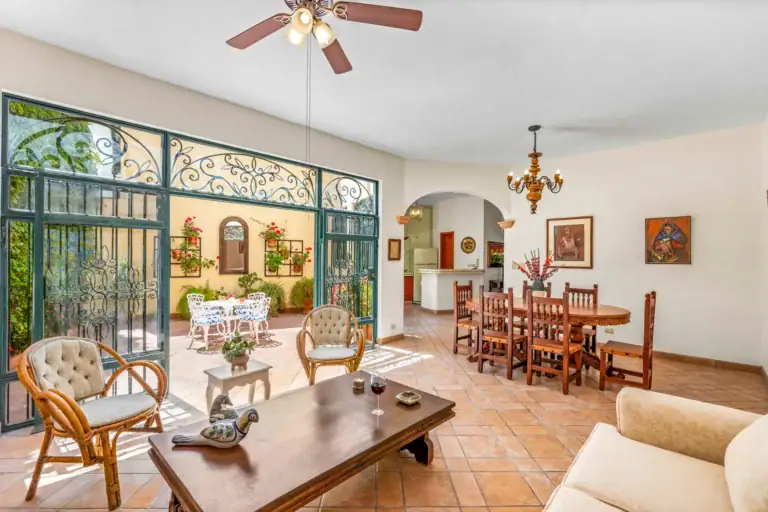 Casa Cul-De-Sac Armonia, living and dining room with a tan sofa, rattan armchairs, a dark wood coffee table and a wrought-iron glass wall to the courtyard, San Miguel de Allende