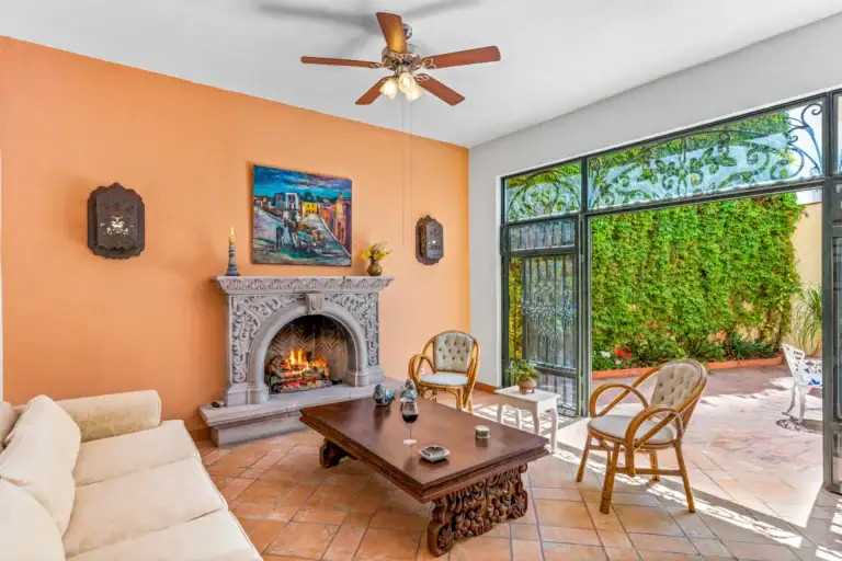 Casa Cul-De-Sac Armonia, living room with an orange wall, a carved stone fireplace with a lit fire, a coffee table and rattan armchairs, San Miguel de Allende