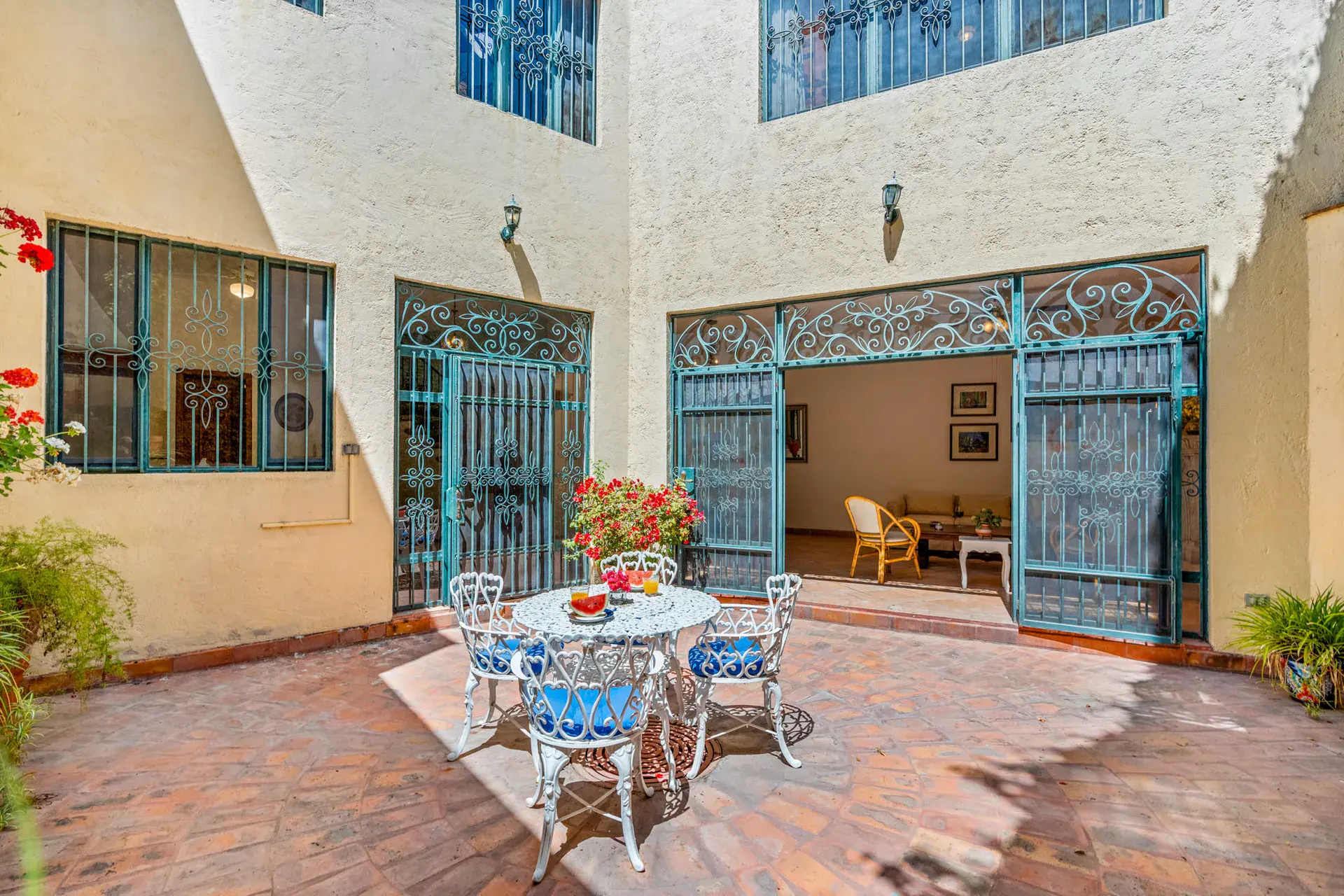 Casa Cul-De-Sac Armonia, courtyard with a brick floor, a white wrought-iron table and chairs, red geraniums and teal iron doors, San Miguel de Allende