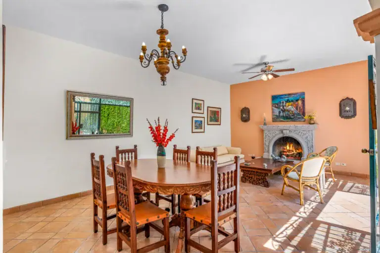 Casa Cul-De-Sac Armonia, dining table with carved wood chairs beside the fireplace and an orange wall, San Miguel de Allende