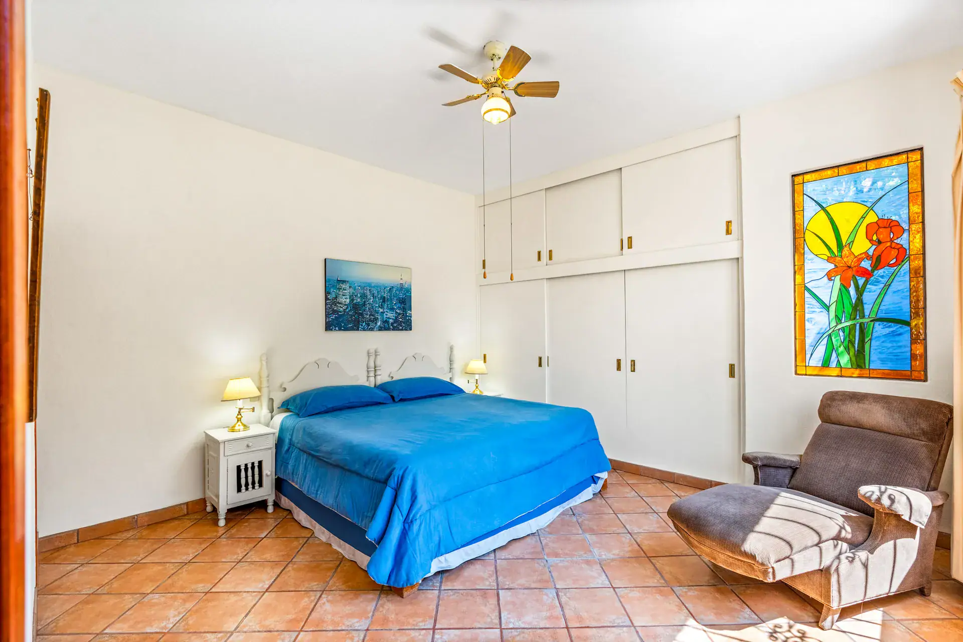 Casa Cul-De-Sac Armonia, bedroom with a blue bedspread, white built-in wardrobes, a stained glass panel and a lounge chair, San Miguel de Allende