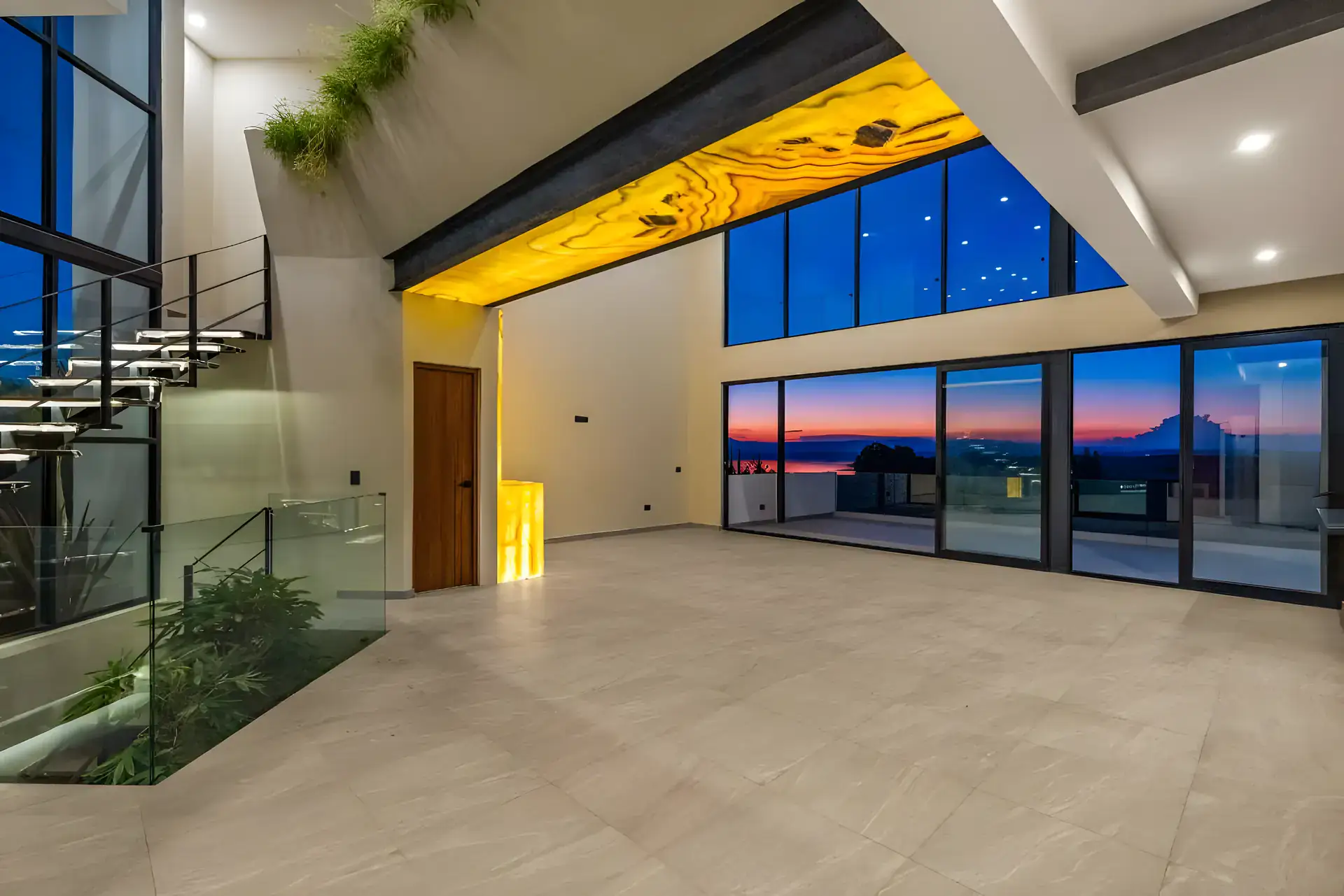 Casa de la Vista, living room at dusk with a glowing onyx wall, a floating stair and glass doors facing a pink sunset sky, San Miguel de Allende