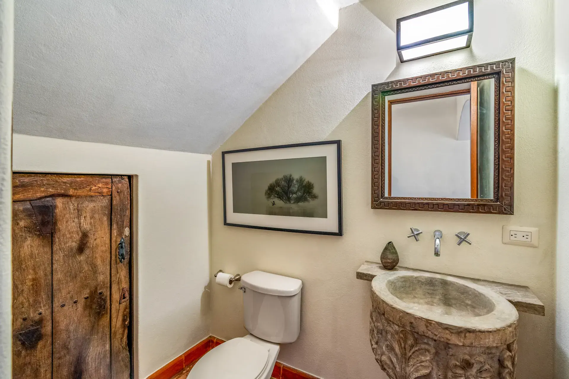 Casa del Artista, powder room with a carved stone sink, a framed mirror, a framed photograph and a rustic wood door, San Miguel de Allende