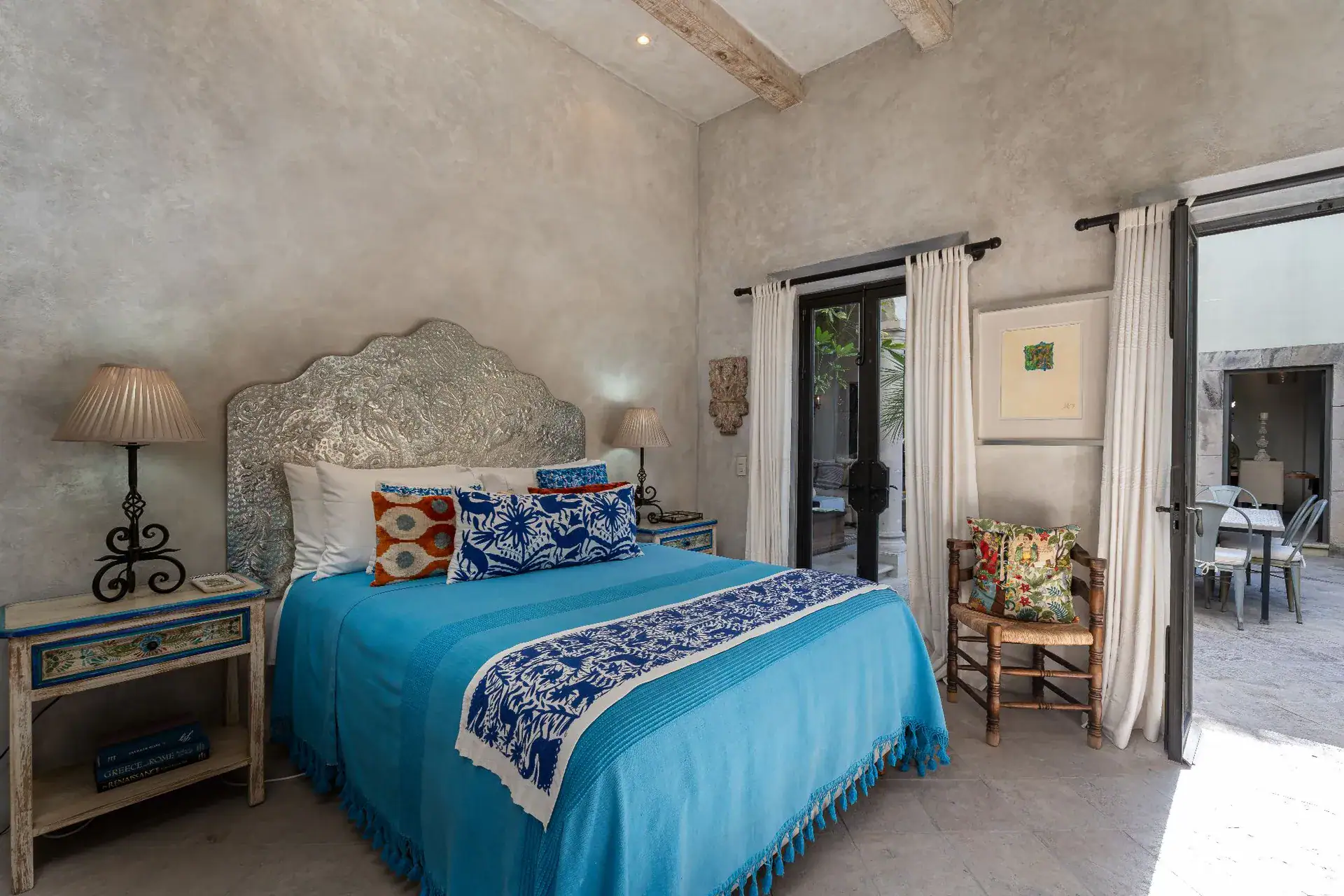 Casa Dos Casas, bedroom with a turquoise bedspread, carved headboard and patterned pillows beside glass doors, San Miguel de Allende