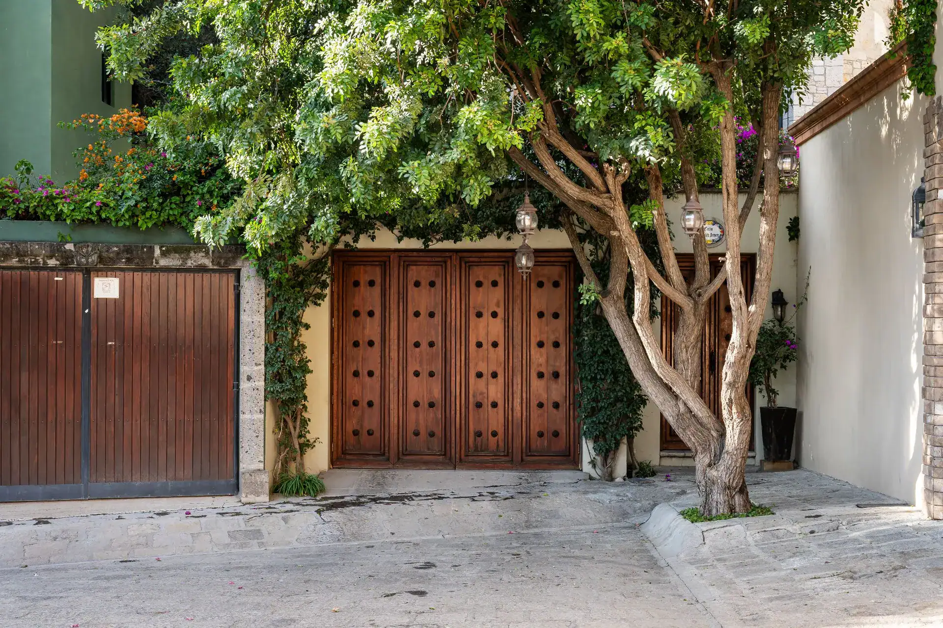 Casa Dos Casas, street entrance with carved wooden double doors and a garage door beneath a leafy tree, San Miguel de Allende