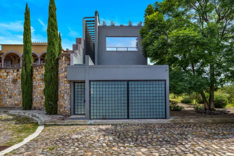 Casa Dos Gatos, modern gray house with a glass-block garage door beside stone walls, cypress trees and a large tree, San Miguel de Allende