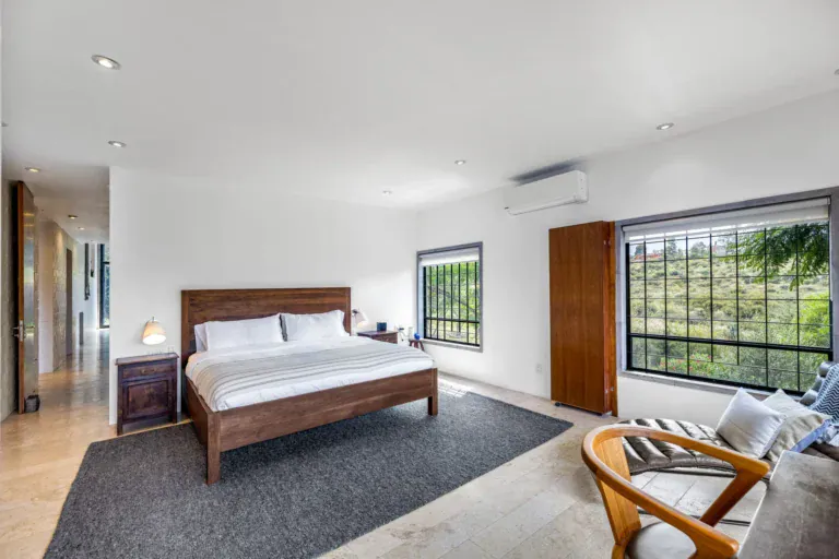 Casa Dos Gatos, bedroom with a wood bed, a gray rug and a large window with a view of the hills, San Miguel de Allende