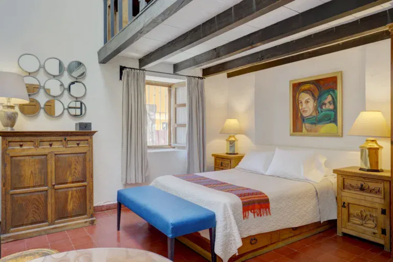 Casa Firenze, bedroom with a white bed, a blue bench, a carved dresser and a loft with beams above, San Miguel de Allende