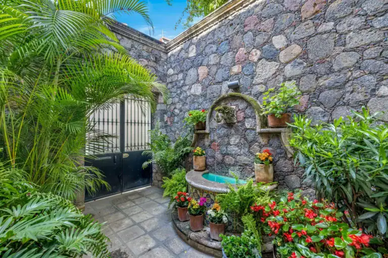 Casa Flora, entry courtyard with a stone wall fountain, a black iron gate and lush plants with red flowers, San Miguel de Allende