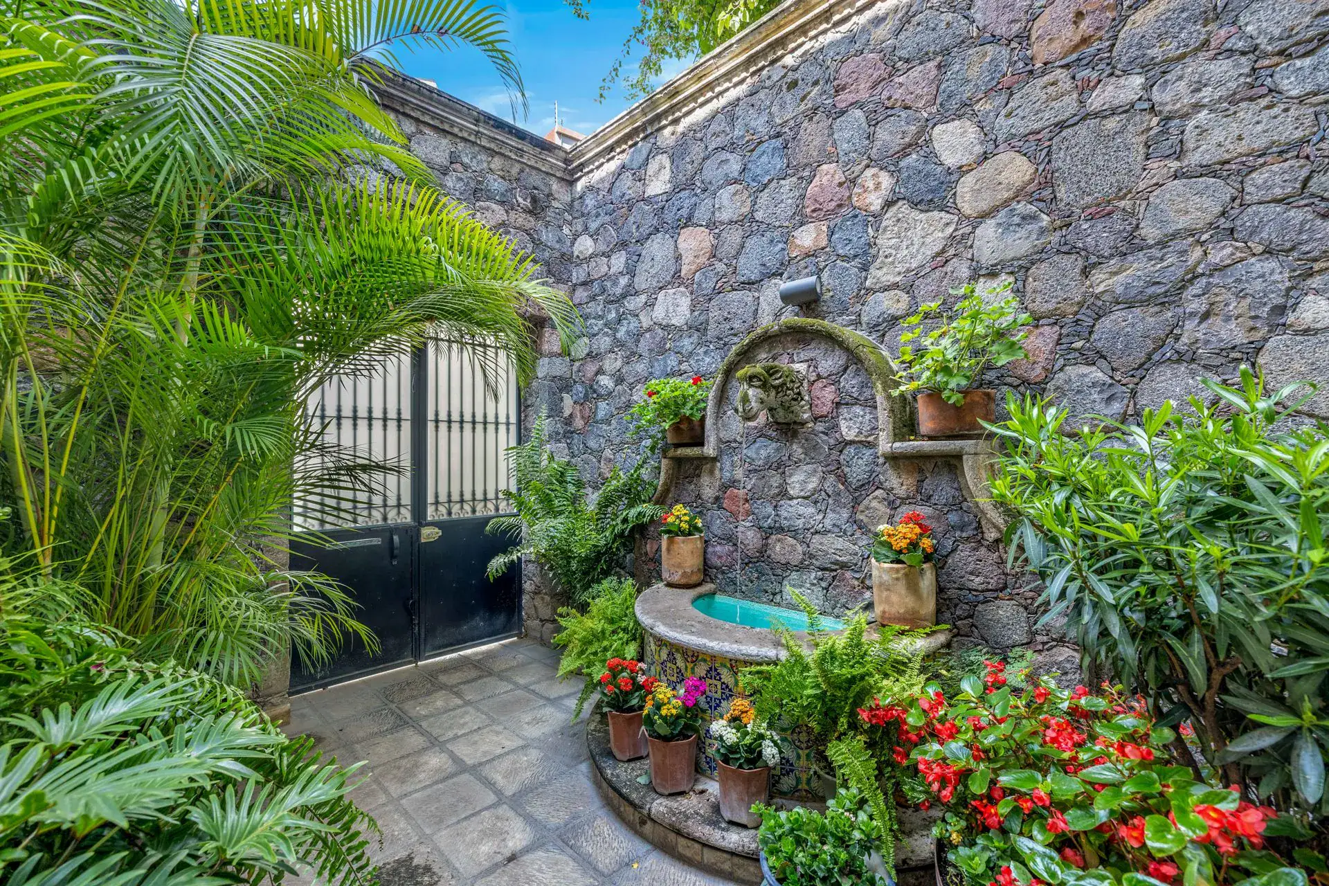 Casa Flora, entry courtyard with a stone wall fountain, a black iron gate and lush plants with red flowers, San Miguel de Allende
