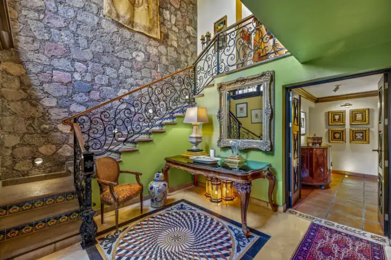 Casa Flora, foyer with a wrought-iron staircase, a stone wall, a green wall and an ornate mirror over a console, San Miguel de Allende